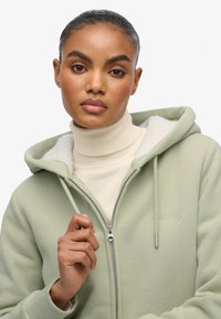 Lichtgroene zip-up hoodie van zacht materiaal, met een kangoeroezak en decoratieve stiksels. Gedragen over een crèmekleurige turtleneck.