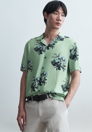 Jeune homme portant une chemise vert clair à manches courtes avec des motifs floraux noirs et un pantalon beige, debout avec une main dans la poche.