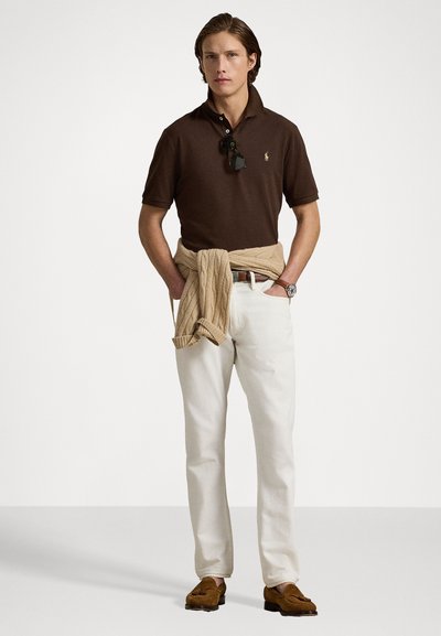 Polo marron, pull beige noué à la taille, pantalon blanc et mocassins marron. Lunettes en caoutchouc et montre-bracelet visibles.