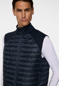 Marinblå pufferväst med hög krage och dragkedja. Har kviltad textur, är ärmlös och har logotypdetalj på kragen.