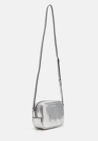 Stříbrná metalická crossbody taška obdélníkového tvaru, s texturovaným povrchem, nastavitelným popruhem a zipovým uzávěrem. Branding viditelný na přední straně.