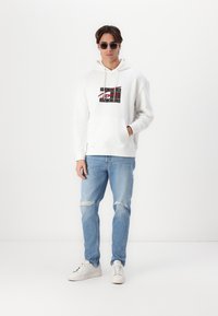 Biela mikina s grafickým logom, predné vrecko a sťahovacie šnúrky. Svetlomodré roztrhnuté džínsy a biele tenisky dopĺňajú outfit.