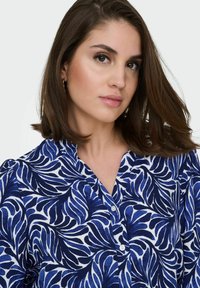 Femme aux cheveux bruns mi-longs portant un chemisier bleu et blanc à motif de feuilles avec des boutons, sur fond blanc.