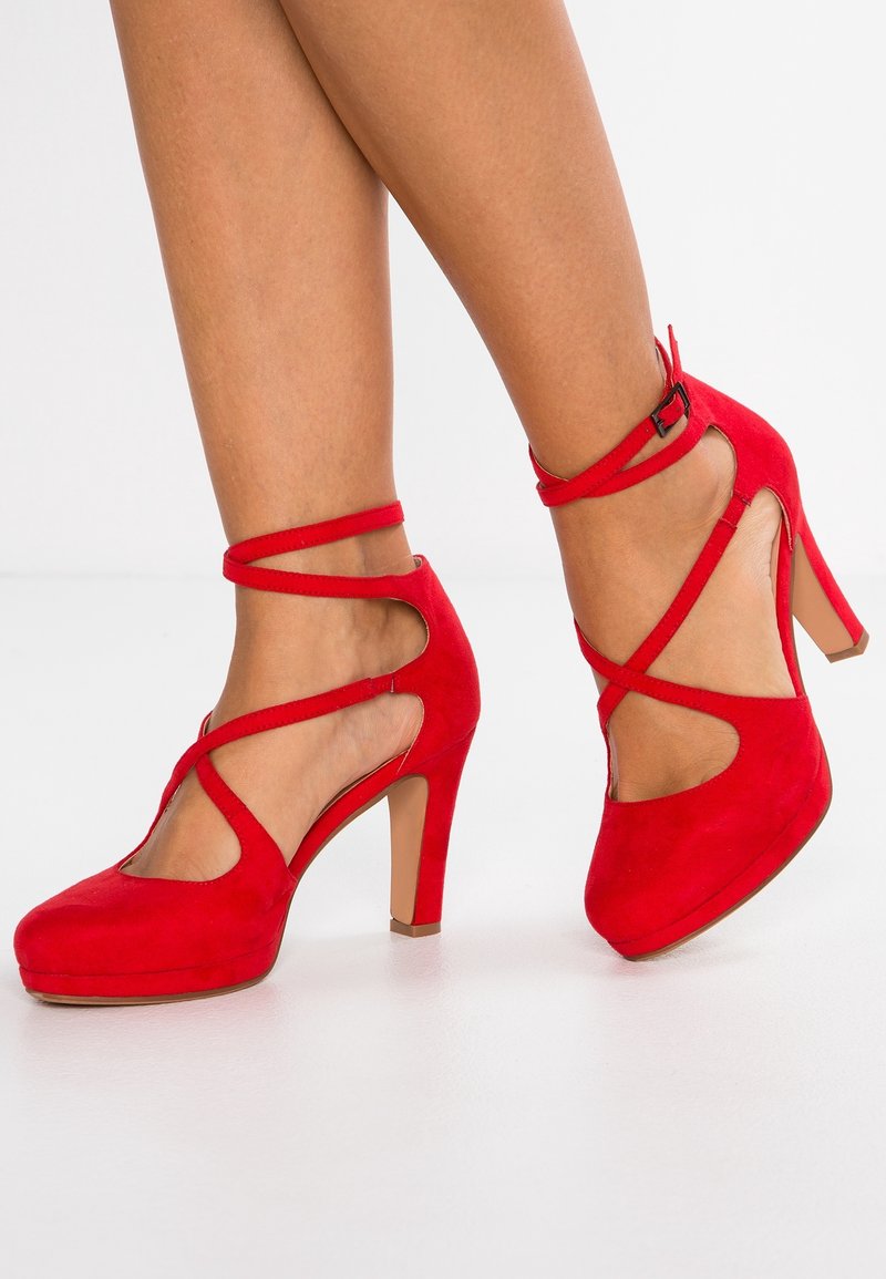 Jambes d'une femme portant des chaussures à talons hauts en daim rouge avec des brides autour de la cheville, sur fond blanc.