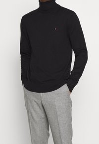 Czarny sweter z golfem wykonany z dzianiny, z małym czerwono-białym logo na piersi. W zestawie z szarymi spodniami o teksturowanej powierzchni.