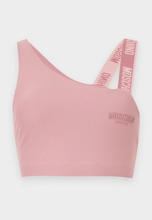 Woman in pink one-shoulder sports bra and matching leggings with branded logos, standing with arms behind back, looking sideways.