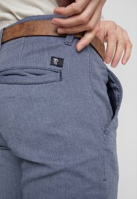 Pantaloni in tessuto blu testurizzato con una vestibilità slim, dotati di una cintura in pelle, tasche frontali e una piccola etichetta del marchio sulla tasca posteriore.