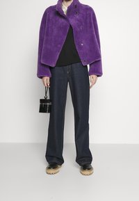 Lila fuskpälsjacka med rund krage och knappstängning, tillsammans med en svart topp, mörk denimbyxor med vida ben och stövlar med tjock sula.