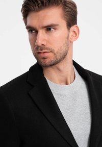 Zwarte blazer met een structuurgrijs geribbeld trui eronder. Scherpe kraag en op maat gemaakte pasvorm, die een modern, minimalistisch ontwerp benadrukt.