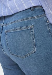 Blue denim jeans feature a back pocket with visible stitching. The fabric has a smooth texture and a mid-rise design.
