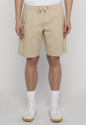 Beige knielange Shorts mit Kordelzugbund, getragen von einer stehenden Person, kombiniert mit weißen Socken und weißen Turnschuhen.