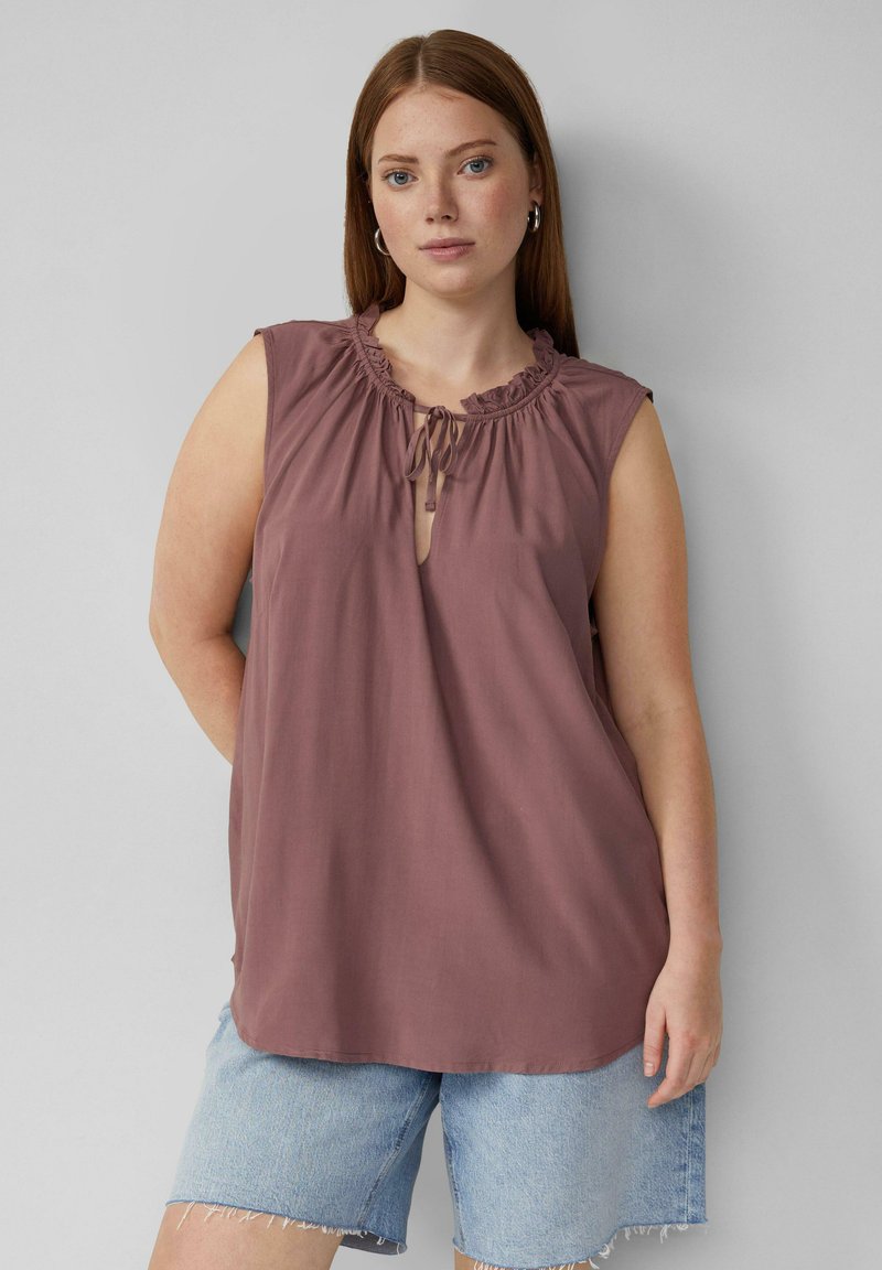 Ärmelloses Oberteil in sanftem Mauve mit Rüschenkragen und Zierschnürung; kombiniert mit hellen Denim-Shorts mit fransigem Saum. Glattes, leichtes Material.
