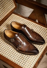 Bruine leren schoenen met een puntige neus, dubbele gesp en een gladde textuur, tentoongesteld op een geweven houten oppervlak.