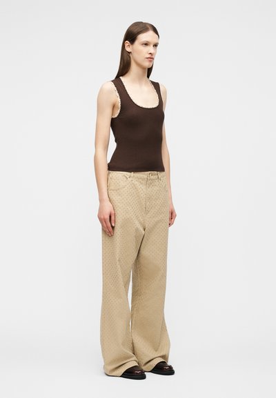 Γυναίκα που φοράει αμάνικη ριγωτή μπλούζα σκούρου καφέ με εκρού περιθώριο, μπεζ παντελόνι με φαρδιά μπάτσα και μικρό πουά σχέδιο, και μαύρα παπούτσια σε λευκό φόντο.