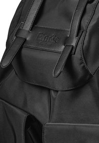 Svart tygryggsäck med en slät yta, vikt klaff och två läderremmar. Varumärkeslogotyp präglad på framsidan. Minimalistisk design.