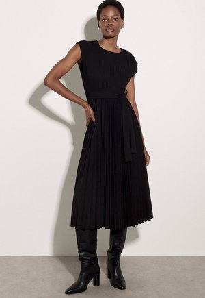 Mujer de pie con un vestido midi negro plisado sin mangas con cinturón en la cintura y botas negras hasta la rodilla con tacón, sobre un fondo liso.