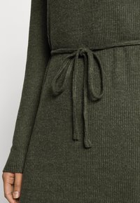 Mörkgrön ribbad klänning med långa ärmar och dragsko i midjan, tillverkad av strukturerat stickat material.