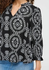 Blusa nera con complessa ricamatura bianca che presenta motivi floreali e geometrici, scollo a V e polsini a volant. Tessuto leggero.