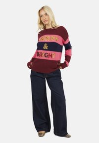 Maglione lavorato a maglia in tonalità di bordeaux, rosa e blu navy con scritta "MERRY & BRIGHT" in paillettes. Abbinato a jeans denim a gamba larga.