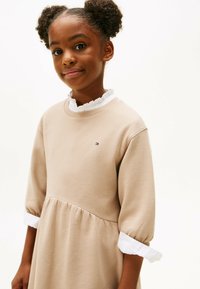 Robe beige à côtes avec un col blanc volanté, des manches 3/4 et une taille froncée. Petit logo sur la poitrine.