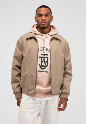 Man die een beige jas draagt over een lichtroze hoodie met grafische tekst, gecombineerd met een witte broek, staand tegen een effen achtergrond.