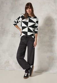 Camicia a maniche lunghe a motivo geometrico in bianco e nero, abbinata a pantaloni neri larghi e sneakers nere con suola marrone, in piedi davanti a uno sfondo testurizzato.