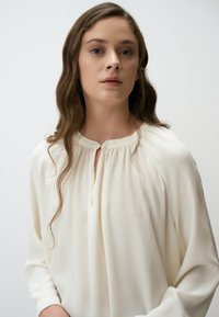 Blusa bianca in tessuto morbido e leggero, con scollatura a chiave, dettagli arricciati alle spalle e maniche lunghe.