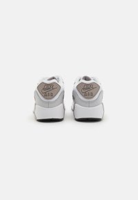 Bijele Nike Air tenisice s teksturiranim sivim logom na peti, ojačanim rupama za vezice i gumenim potplatom. Glatki kožni i mrežasti materijali.