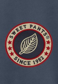 Patch circulaire brodé avec un design de feuille, bord rouge, texte blanc disant "SWEET PANTS®" et "SINCE 1982," avec des étoiles. Fond sombre.