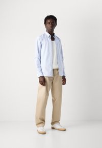 Lyseblå stripete skjorte med knapper over en hvit tanktop, paret med beige bukser og hvite sneakers med brune detaljer. Glatt stoff og avslappet passform.