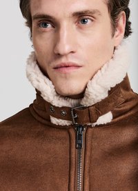 Giacca marrone in ecopelle con un morbido colletto in shearling beige. Dotata di chiusura con zip e bottoni a pressione. Superficie texturizzata e design strutturato.