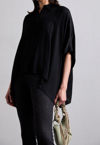 Blusa negra de corte holgado con cuello y aberturas laterales, combinada con jeans oscuros y un pequeño bolso tejido verde con asas texturizadas.
