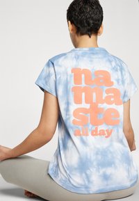 Zila un baltā krekliņa ar saites krāsojumu, īsām piedurknēm, ar lieliem oranžiem uzrakstiem "namaste all day" aizmugurē. Mīksta auduma, brīvas piegriezuma.