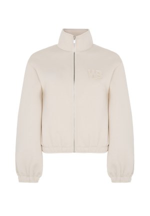 Beige ritsjack met een hoge kraag, voorzien van elastische manchetten en een elastische tailleband, en een geborduurd "WB" logo op de borst. Soepele textuur.