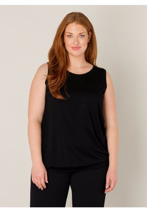 Smiling woman with long red hair wearing a sleeveless black top and black pants, standing against a plain light background.