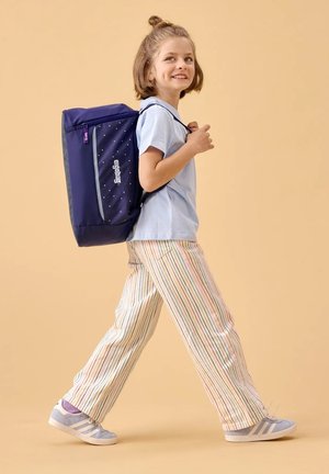 Navy blue backpack with white speckles, adjustable straps, and a front pocket. Model in light blue shirt and striped trousers, with gray shoes.