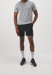 Šedé texturované sportovní tričko s vzorem chevron, spárované s černými šortkami. Nošeno s bílými ponožkami a černými běžeckými botami s šedými akcenty.