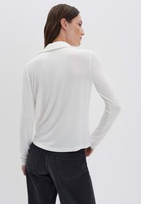 Donna con lunghi capelli castani, vestita con una maglietta bianca a maniche lunghe e jeans neri, in piedi con una mano sulla vita, girata di spalle.