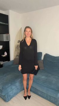 Sort langermet kjole med v-hals i glatt tekstur, kombinert med svarte spisse hæler. Holder en clutch, står på en blå sofa.