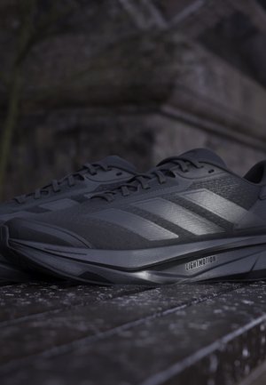 Chaussures de course athlétiques noires avec des bandes réfléchissantes et un amorti « Lightmotion » sur une surface texturée sombre en faible luminosité.