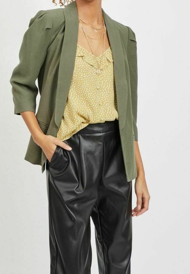 Femme portant un blazer vert olive, un chemisier jaune à pois, des colliers en or superposés et un pantalon en cuir noir taille haute, une main dans la poche.