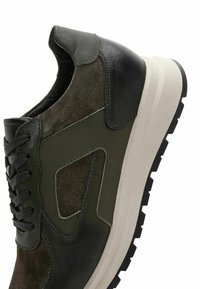 Zapatilla verde oscuro que combina cuero liso y gamuza, con una suela blanca gruesa y una suela texturizada negra. Diseño con cordones.