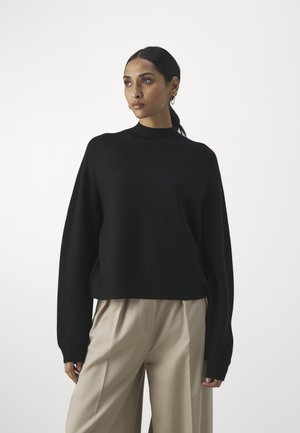 Schwarzer, übergroßer Pullover mit hohem Kragen und langen Ärmeln, aus einem glatten Stoff; kombiniert mit hellbeigen, weit geschnittenen Hosen.