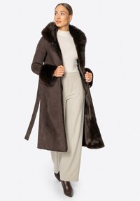 Brązowy płaszcz z futra syntetycznego z szerokim kołnierzem i mankietami, z paskiem do związania i bocznymi rozcięciami, noszony na jasny sweter i beżowe spodnie.