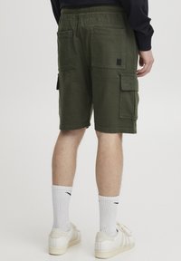 Oliven grønne cargo shortser med elastisk talje, to store sidelommer og en stofmærke, parret med hvide ribbede sokker og sneakers.