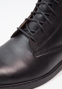 Stivale alla caviglia in pelle nera con texture liscia, caratterizzato da una stringatura aderente, dettagli di cucitura sottili e una punta arrotondata. Suola interamente nera.