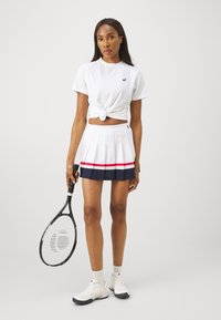 T-shirt atletico bianco annodato in vita, abbinato a una gonna plissettata bianca e blu navy con strisce rosse. Tiene in mano una racchetta da tennis nera.