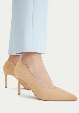 Nudowe zamszowe szpilki z ostrym noskiem noszone z jasnoniebieskimi krótkimi spodniami na jednolitym tle.