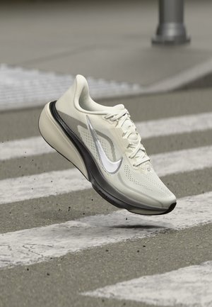 Lyse farvede Nike løbesko med sort sål svævende over et fodgængerfelt på en grå asfaltvej.
