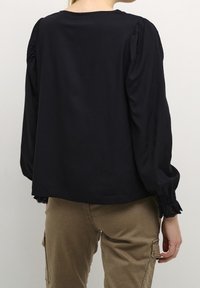 Blusa nera a maniche lunghe con spalle arricciate, bordo in pizzo sui polsini e vestibilità morbida. Indossata con pantaloni cargo color sabbia.